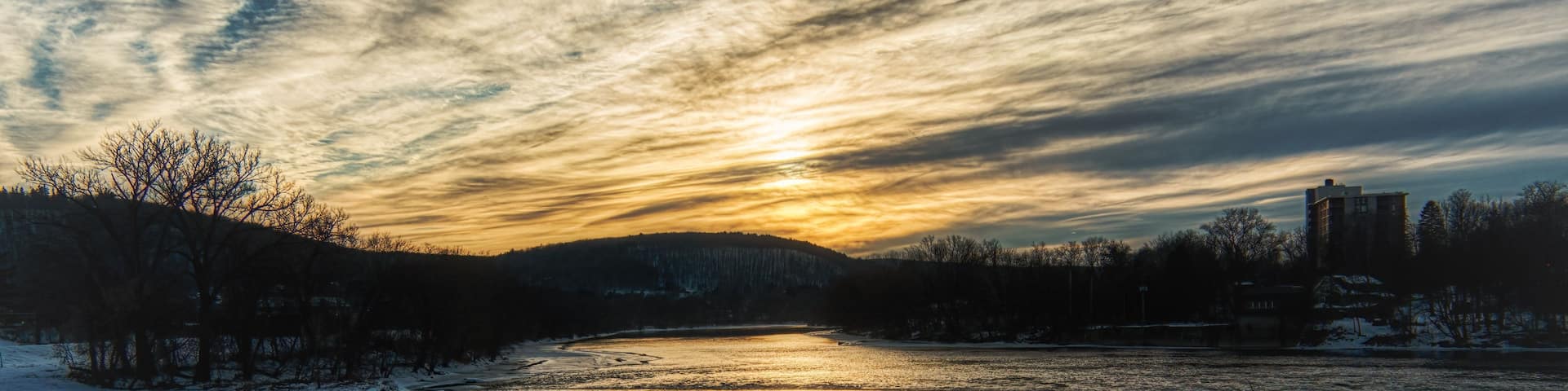 A frozen river with a partially frozen surface, surrounded by trees and buildings under a sunset sky.