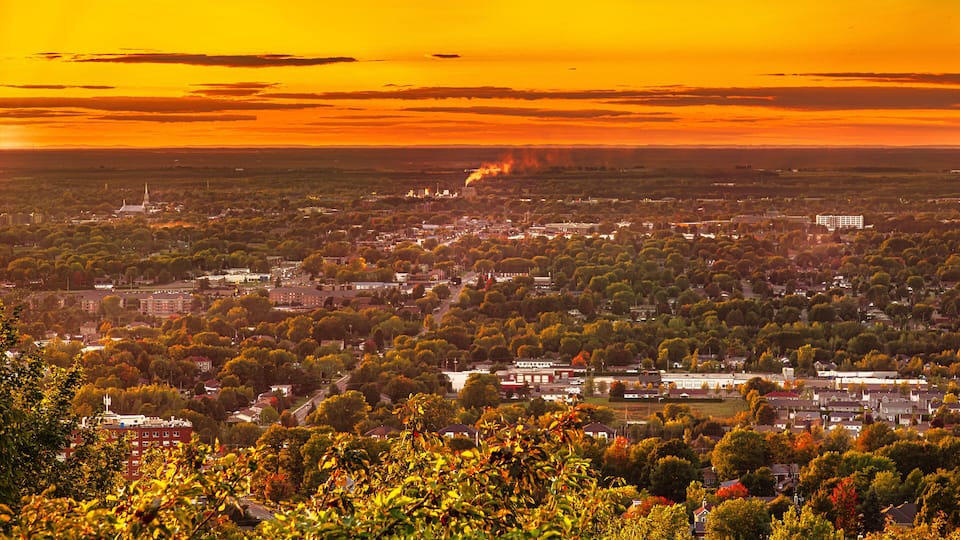 A cityscape at sunset with a prominent church spire, dense tree coverage, and a factory emitting smoke.