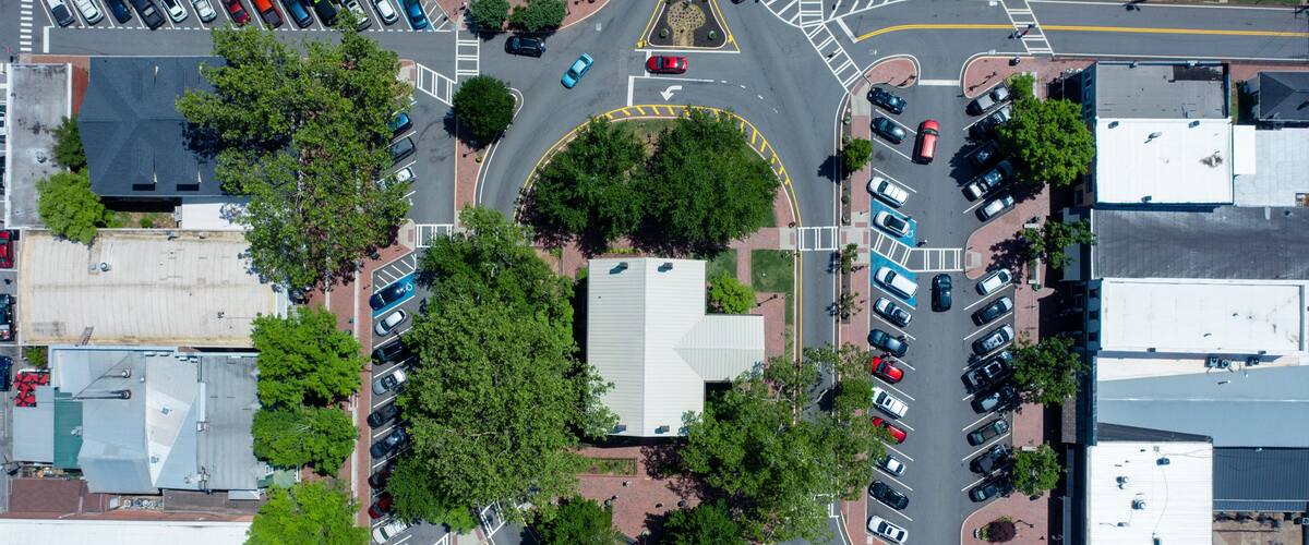 An aerial view of a roundabout with multiple lanes, surrounded by parking lots and buildings.