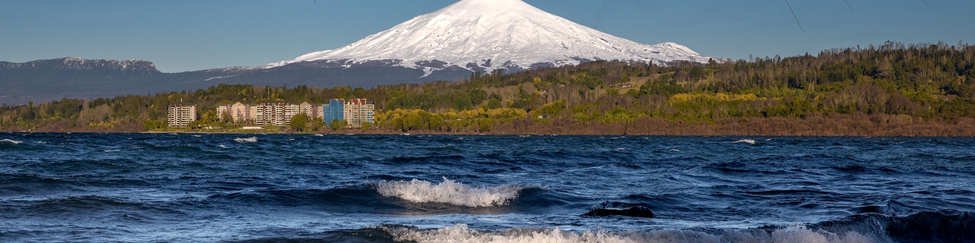 Montanha nevada, um corpo de água com ondas e uma área florestada com edifícios ao fundo.