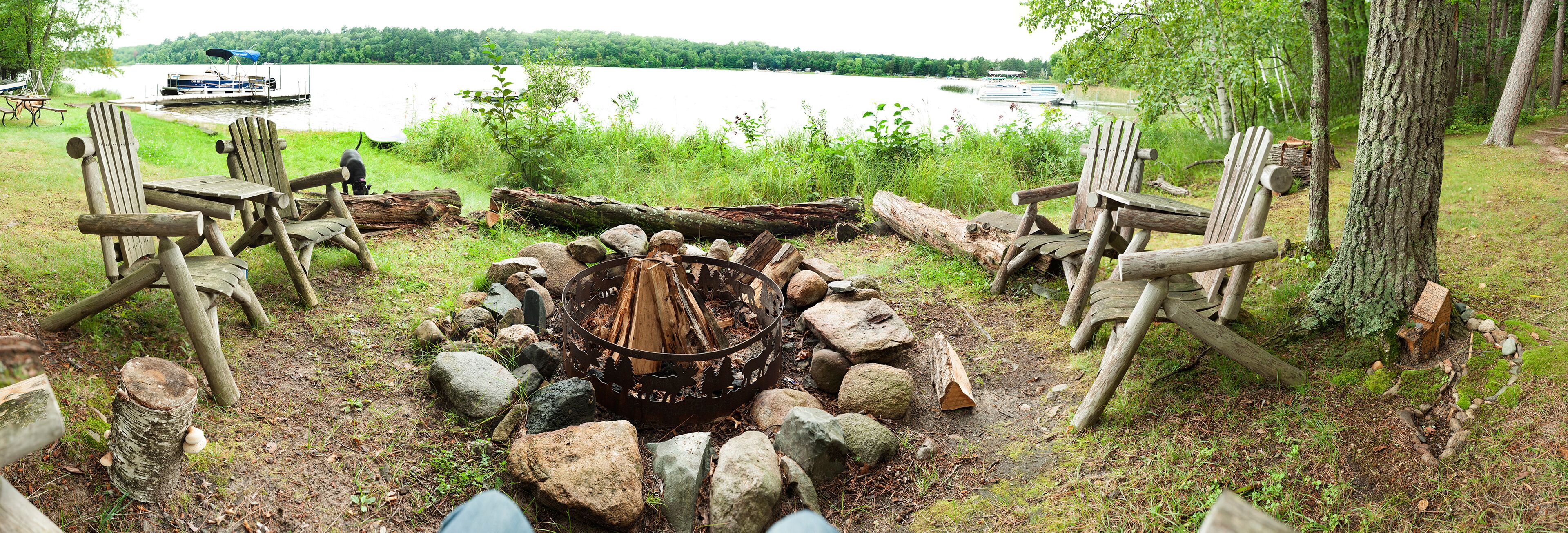 A lakeside campfire area with Adirondack chairs and a fire pit surrounded by rocks.