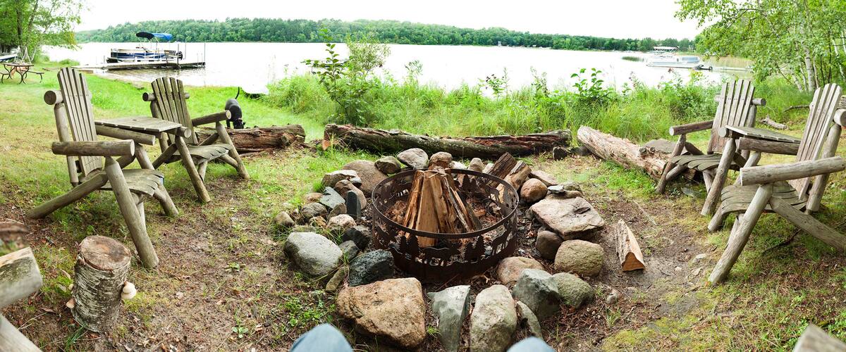 A lakeside campfire area with Adirondack chairs and a fire pit surrounded by rocks.