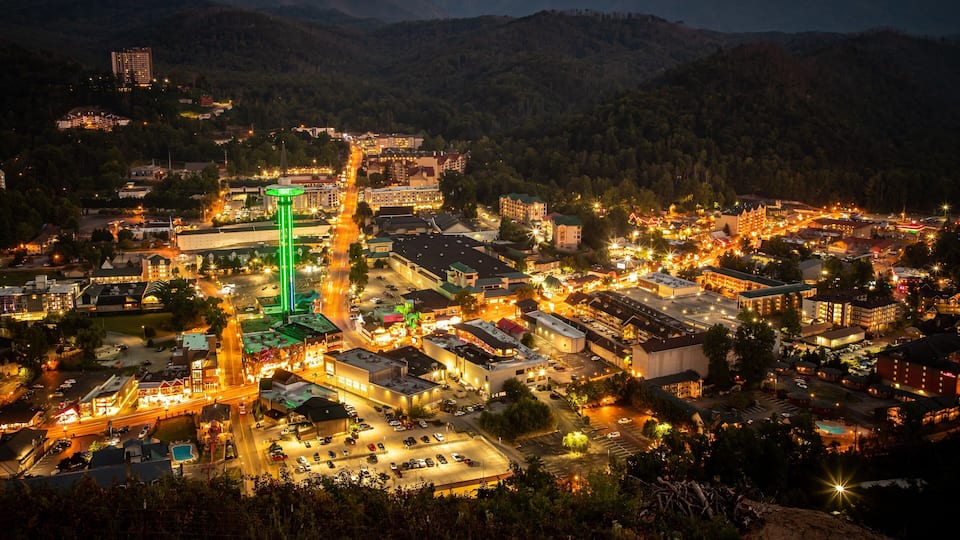 A town at night with a prominent green-lit tower, surrounded by buildings and a mountainous backdrop.