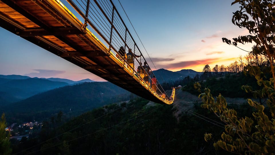 A suspension bridge at sunset with mountains in the background.