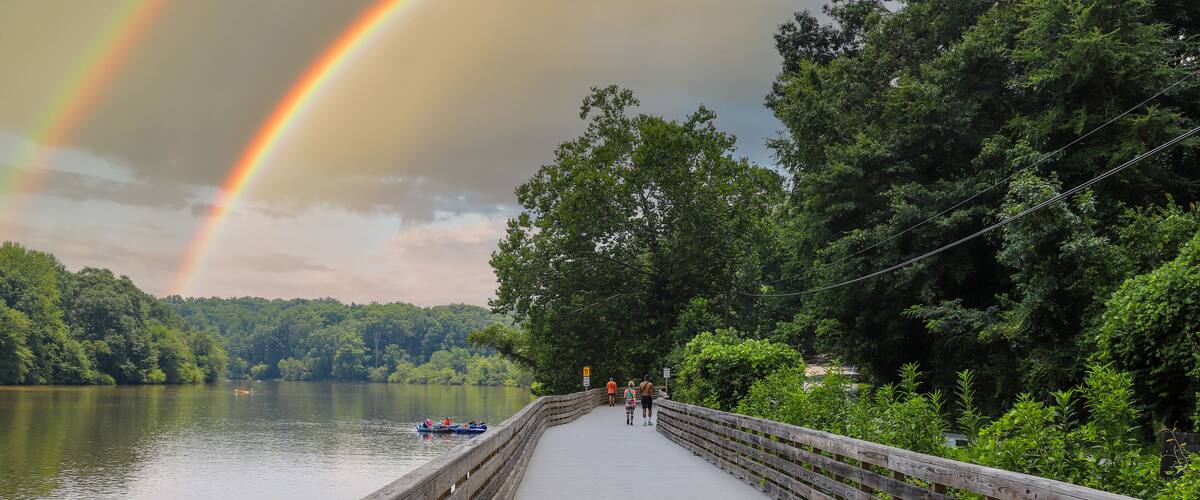 people walking down the boardwalk along the Chattahoochee river with vast silky brown river water surrounded by lush green trees at Roswell Riverwalk Boardwalk in Roswell Georgia USA