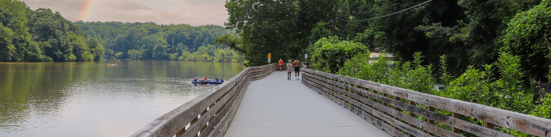 people walking down the boardwalk along the Chattahoochee river with vast silky brown river water surrounded by lush green trees at Roswell Riverwalk Boardwalk in Roswell Georgia USA