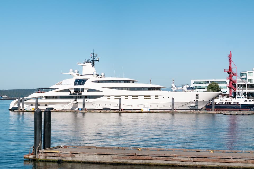 Een grote witte jacht afgemeerd in een jachthaven, met een rode kraan en moderne gebouwen op de achtergrond.