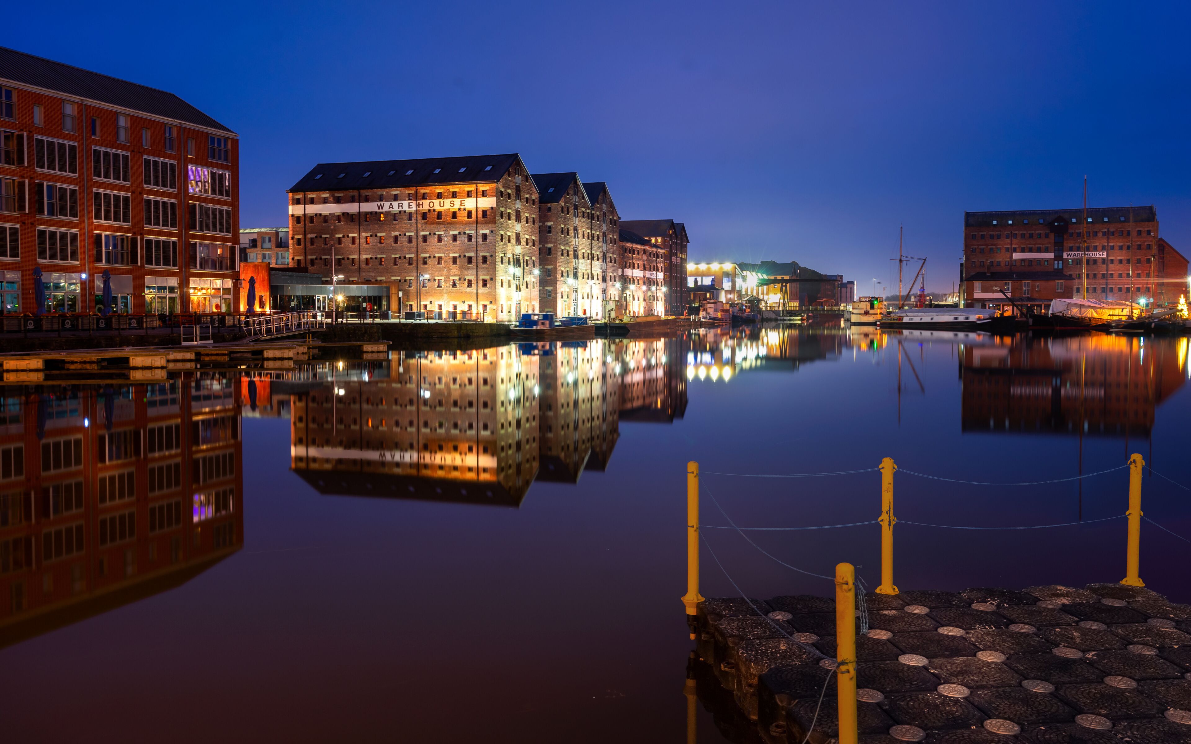 A waterfront cityscape at dusk with illuminated buildings, including a warehouse, reflecting in the calm water.