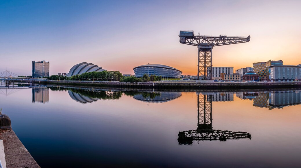 A city skyline with a large Ferris wheel, modern buildings, and a calm river reflecting the structures.