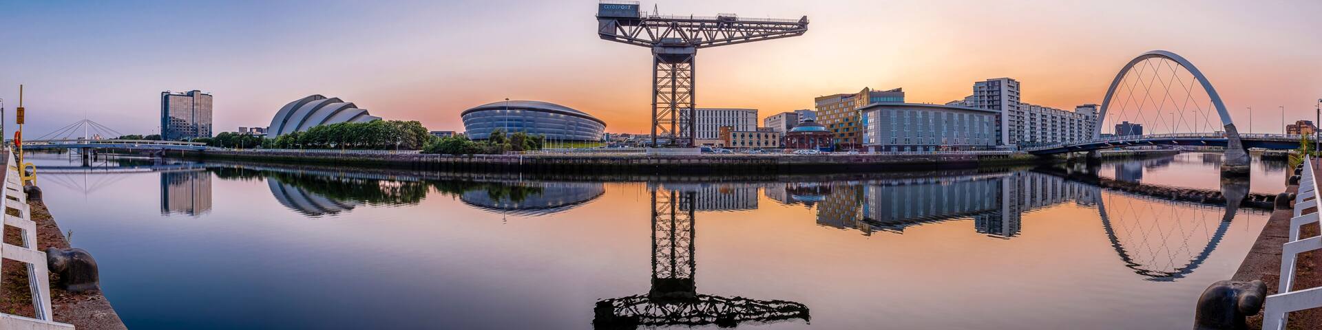 A city skyline with a large Ferris wheel, modern buildings, and a calm river reflecting the structures.