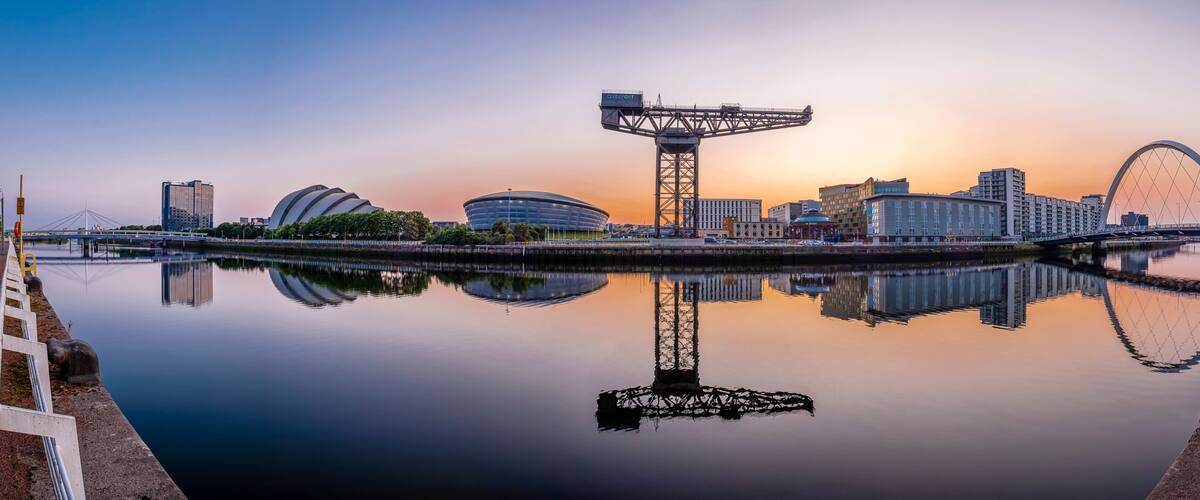 A city skyline with a large Ferris wheel, modern buildings, and a calm river reflecting the structures.