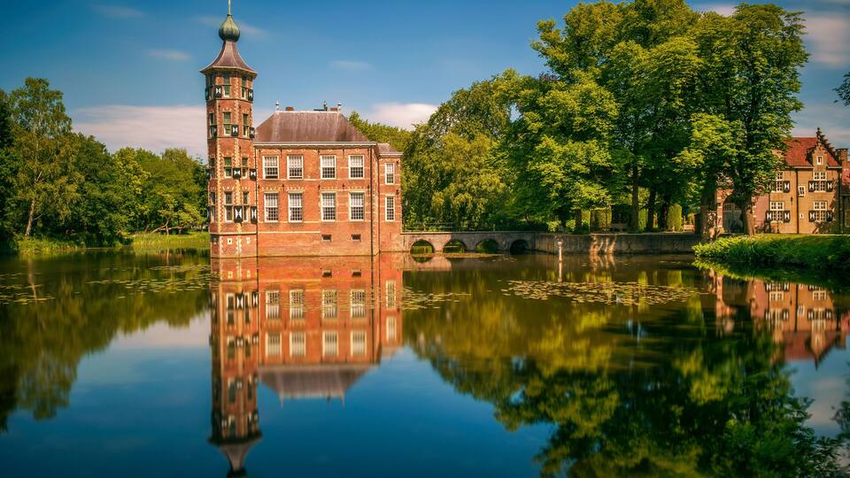 A historic brick building with a tower, reflecting in a calm lake, surrounded by lush trees.