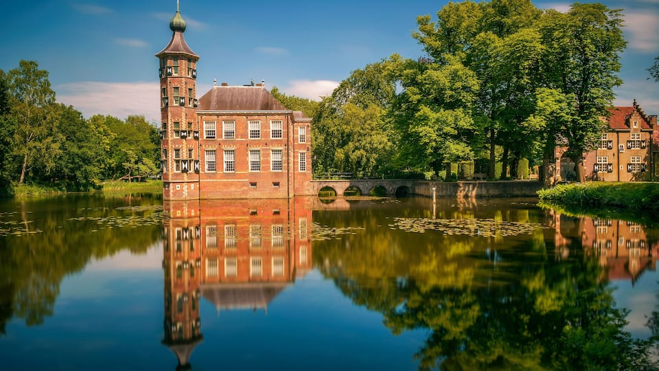 A historic brick building with a tower, reflecting in a calm lake surrounded by lush trees.