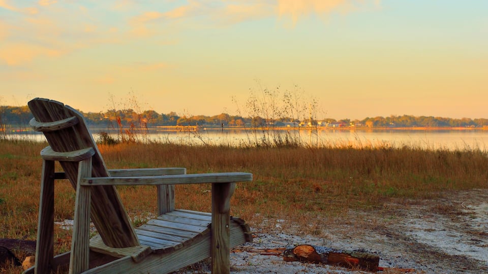 A wooden Adirondack chair on a sandy beach with a lake and trees in the background.