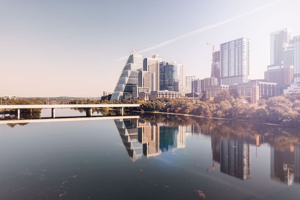 A city skyline reflected in calm water, with a prominent modern building and a bridge in the foreground.