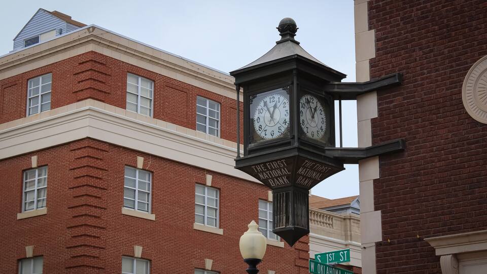 A clock tower with two clocks, a street sign reading "1st St Oklahoma Ave", and a brick building in the background.