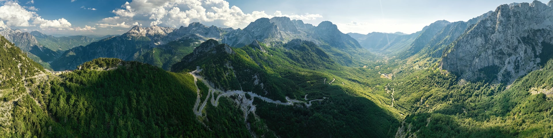A panoramic, aerial view of the winding road into the Theth Valley. Green valleys, blue sky with clouds, steep rocks, remnants of snow. Theth national park, Albanian Alps, Albania.