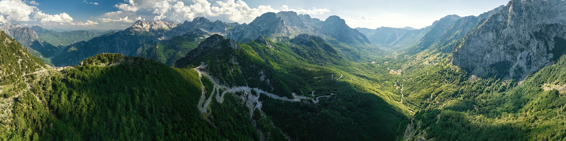 A panoramic, aerial view of the winding road into the Theth Valley. Green valleys, blue sky with clouds, steep rocks, remnants of snow. Theth national park, Albanian Alps, Albania.