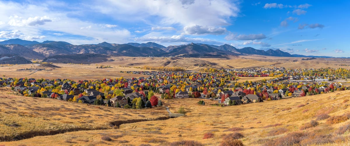 A residential area with houses surrounded by autumn-colored trees and mountains in the background.