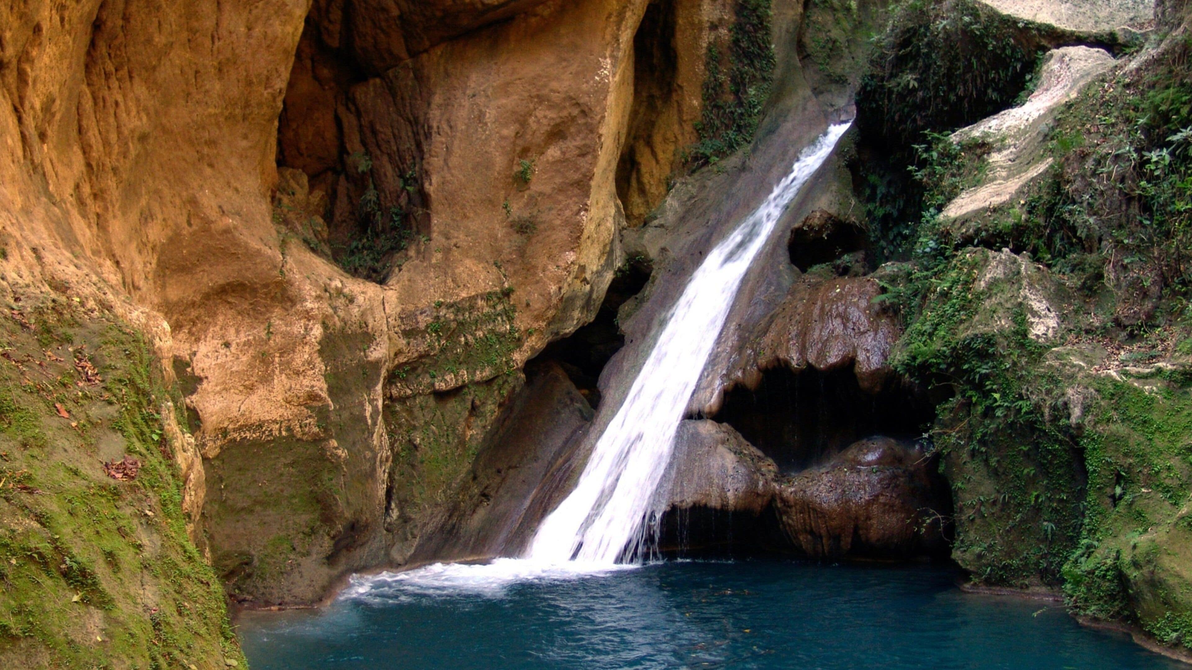 A waterfall cascading down a rocky cliff into a pool of water surrounded by lush greenery.