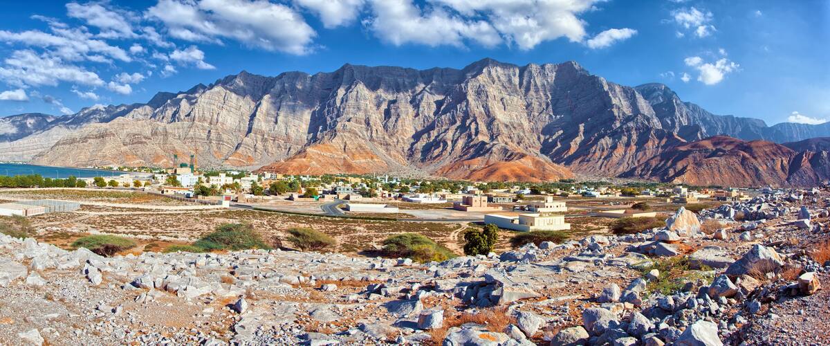 A coastal town with rocky terrain, a large mountain range in the background, and a clear blue sea.