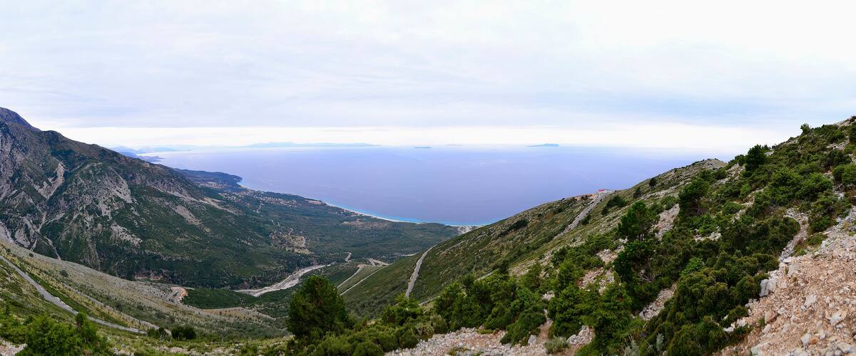 A mountainous landscape with a winding road, dense greenery, and a distant view of the sea.