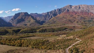 Autumn fields near Llugaj open toward the towering Albanian Alps and the winding mountain road to Bajram Curri.