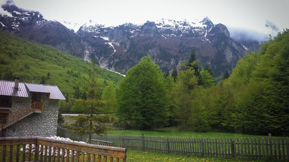 A stone house with a wooden deck overlooking a valley with a wooden fence and a field of yellow flowers.