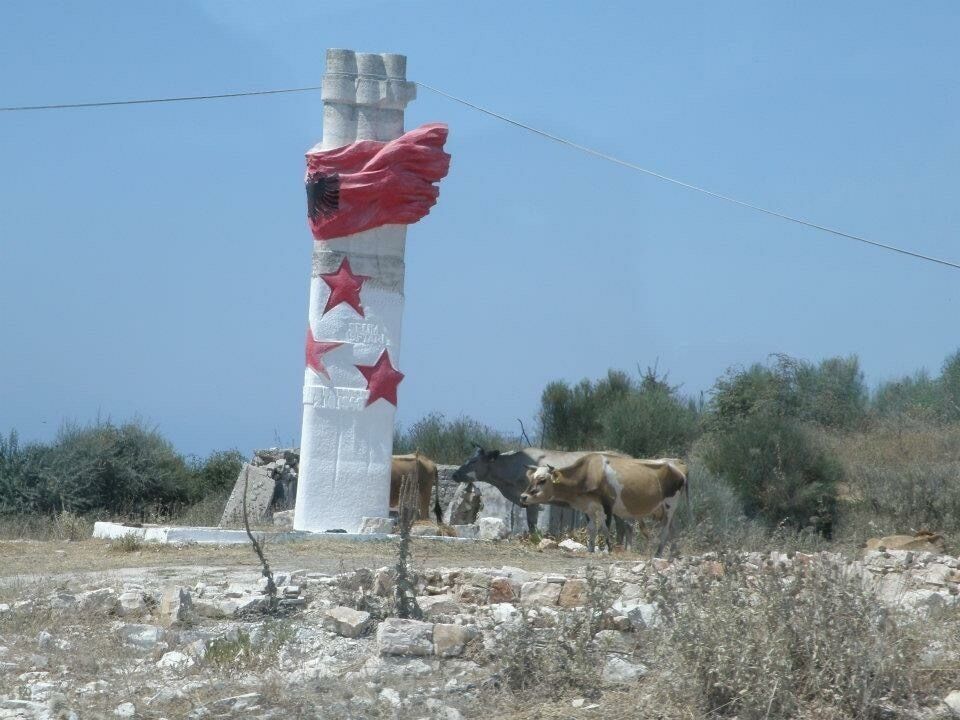 A white column with red stars and a red flag, surrounded by cows and dry vegetation.
