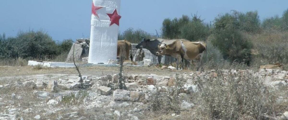 A white column with red stars and a red flag, surrounded by cows and dry vegetation.