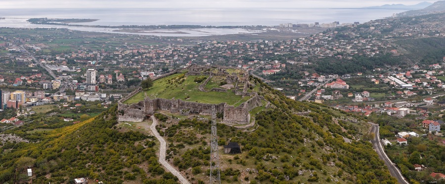 Aerial view of Lezhë Castle overlooking the city, surrounding hills, and the Adriatic coastline under a soft cloudy sky.
