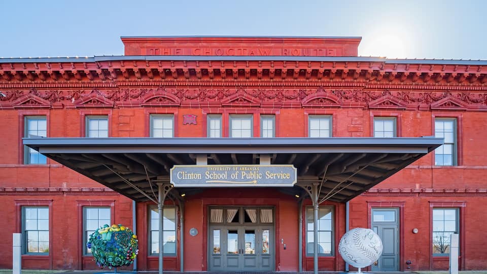 A red brick building with a sign reading "Clinton School of Public Service" and a baseball statue.