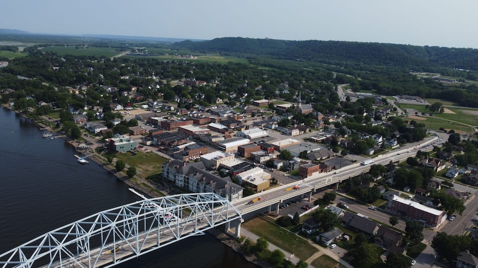 A river with a bridge, a town with buildings, and green hills in the background.