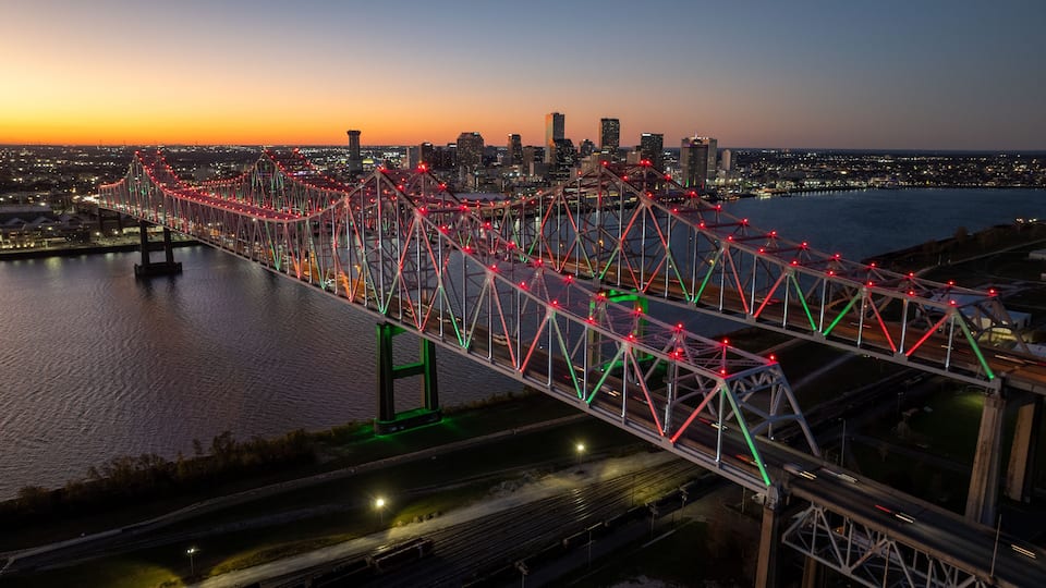 A brightly lit bridge with red and green lights against a city skyline at dusk.