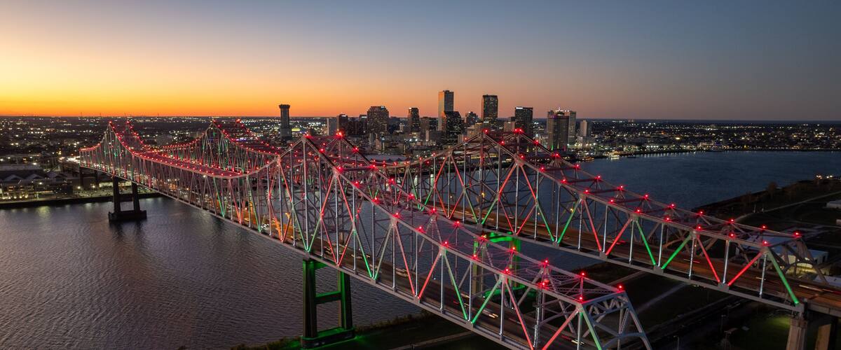 A brightly lit bridge with red and green lights against a city skyline at dusk.
