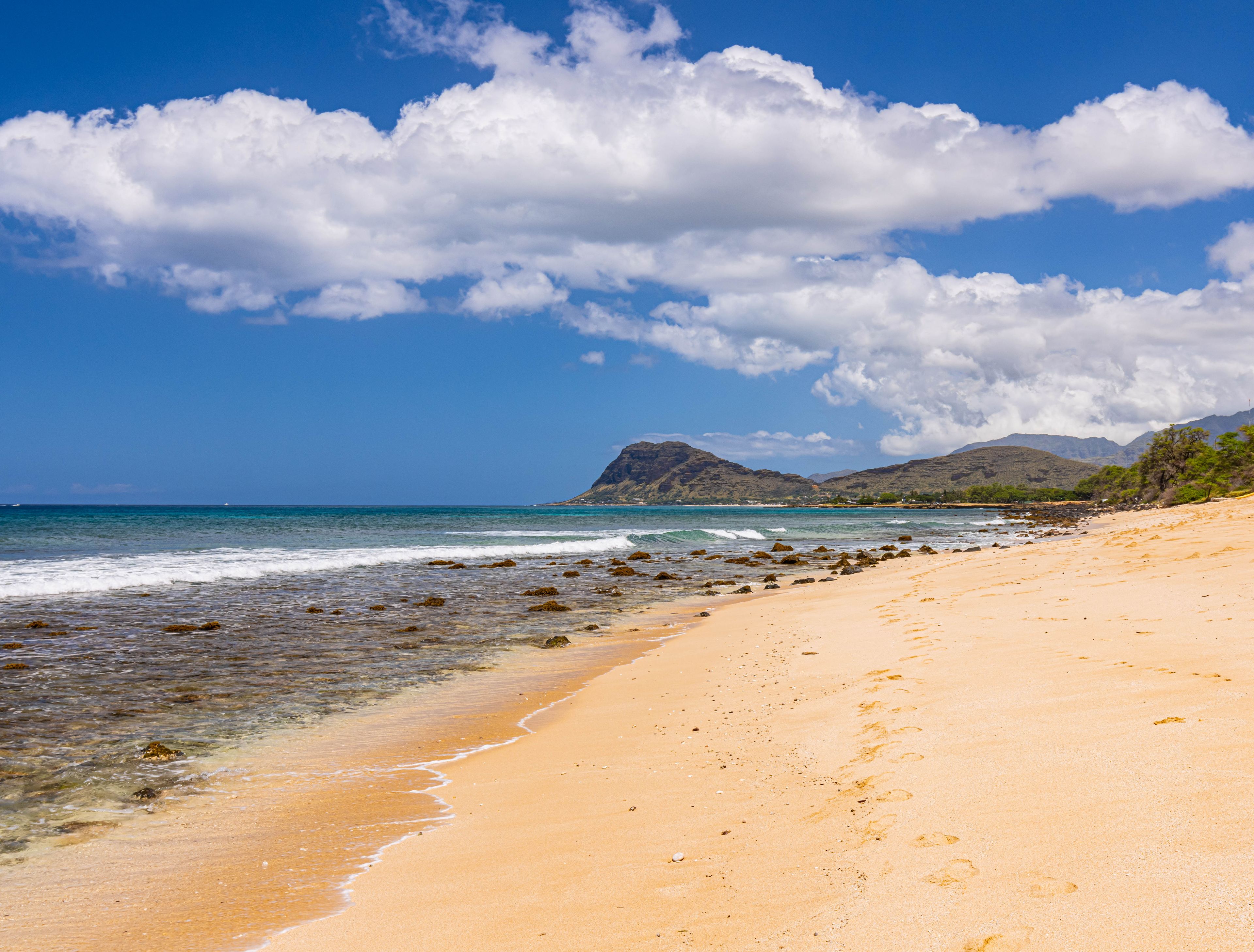 A beach with clear blue water, sandy shore, and distant mountains under a partly cloudy sky.