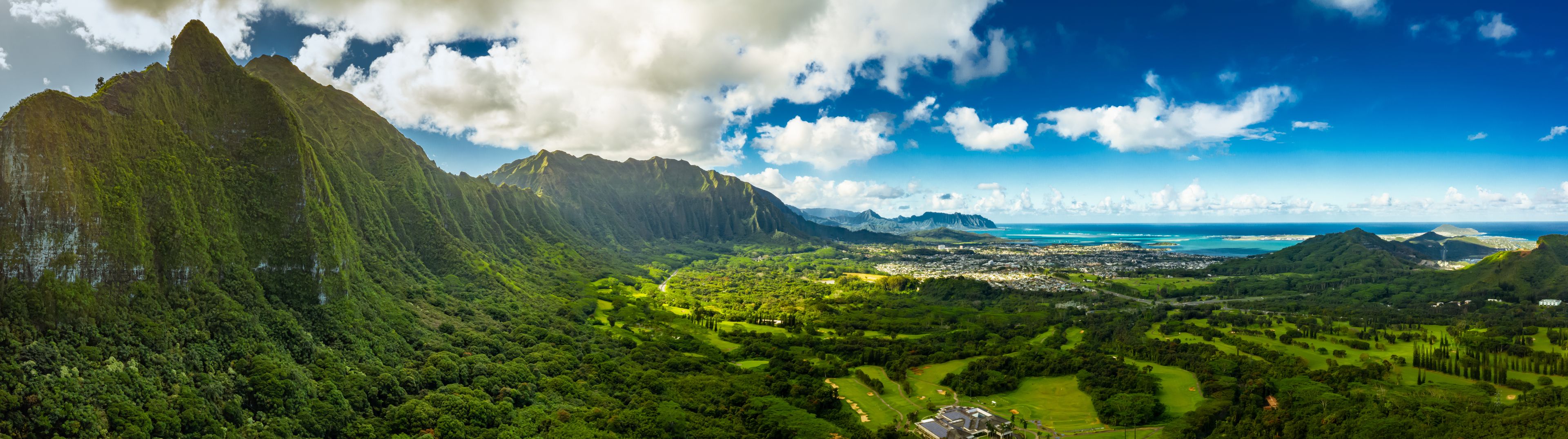 A Panoramic aerial image from the Pali Lookout on the island of Oahu in Hawaii.  With a bright green rainforest, vertical cliffs and vivid blue skies.