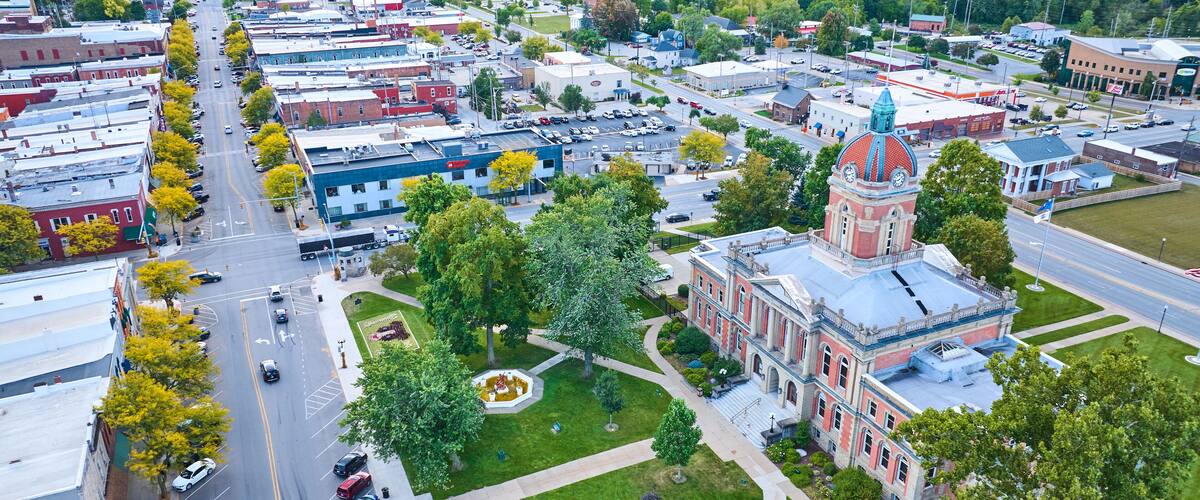 An aerial view of a town with a prominent clock tower, a central green space, and a main street lined with trees and parked cars.