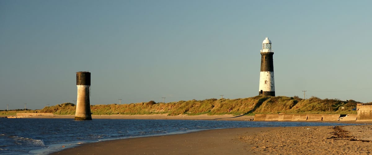 A coastal scene with two lighthouses, a sandy beach, and a grassy hill in the background.