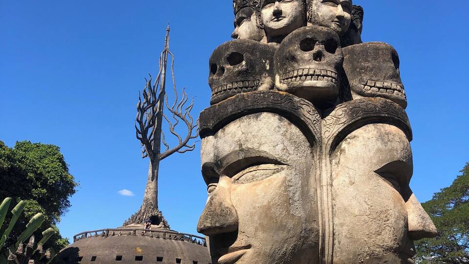 A large stone statue with multiple faces and a tree in the background.