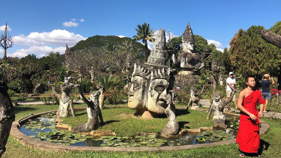 A large stone Buddha statue surrounded by a pond with lily pads, set in a lush garden with palm trees and other greenery.