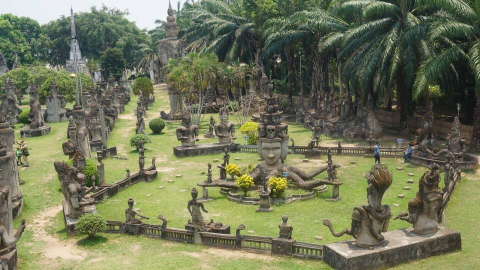 A temple complex with numerous statues and a central Buddha statue surrounded by palm trees.