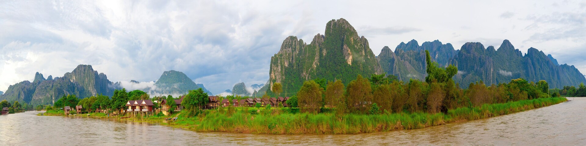 Um rio com casas sobre palafitas, cercado por vegetação exuberante e montanhas imponentes.