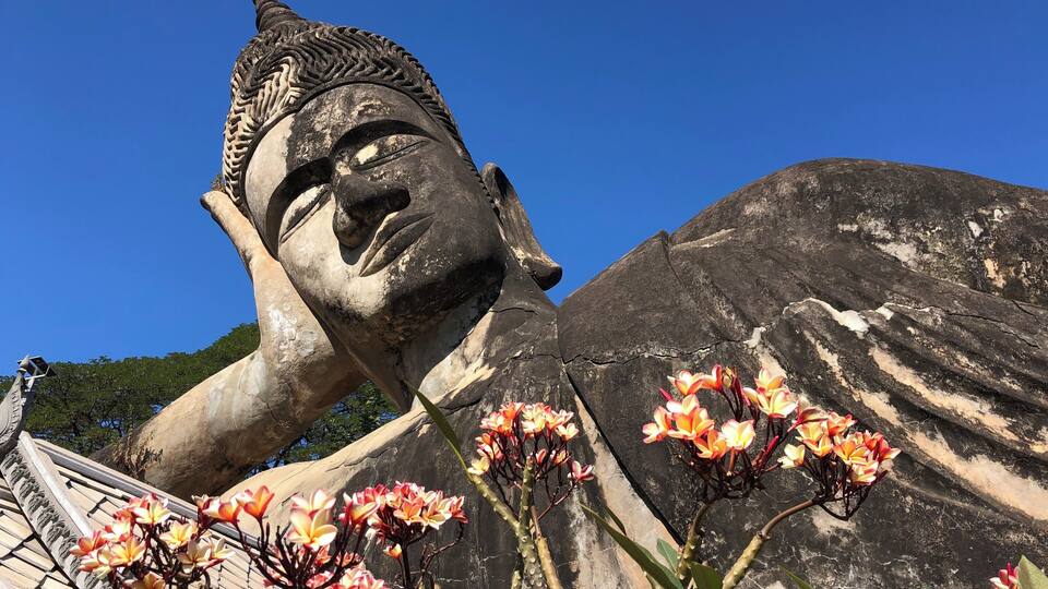A large Buddha statue with intricate carvings and a serene expression, surrounded by blooming flowers.
