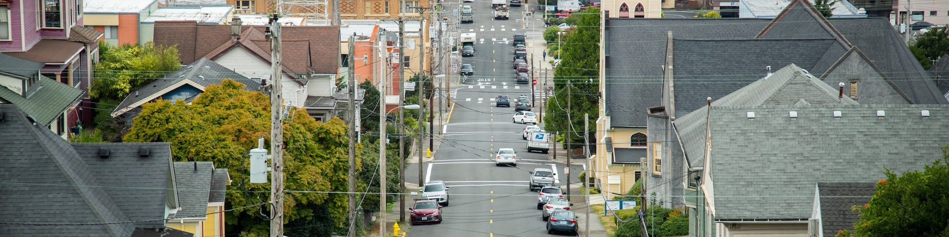 A coastal street lined with houses and buildings, with cars parked along the road and a view of the water in the distance.