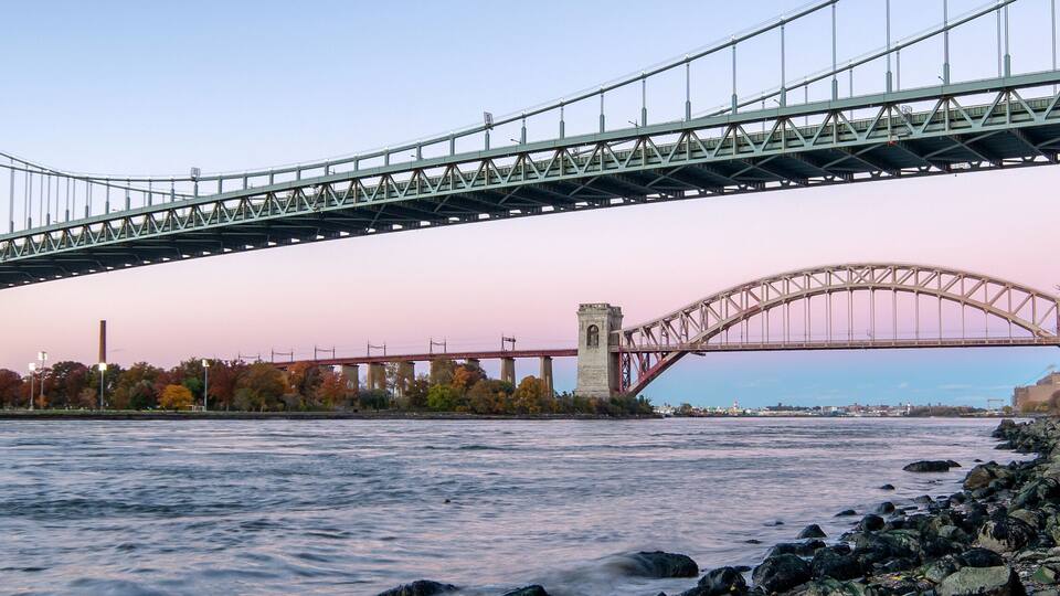 A large suspension bridge spanning a river, with a stone wall and autumn foliage along the riverbank.