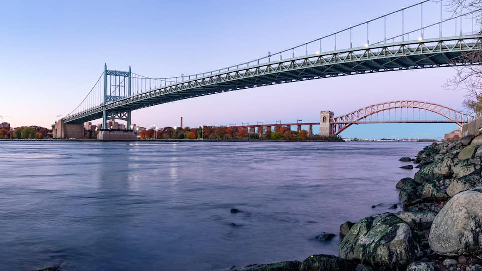 A large suspension bridge spanning a river, with a stone embankment on the right and autumn-colored trees in the background.