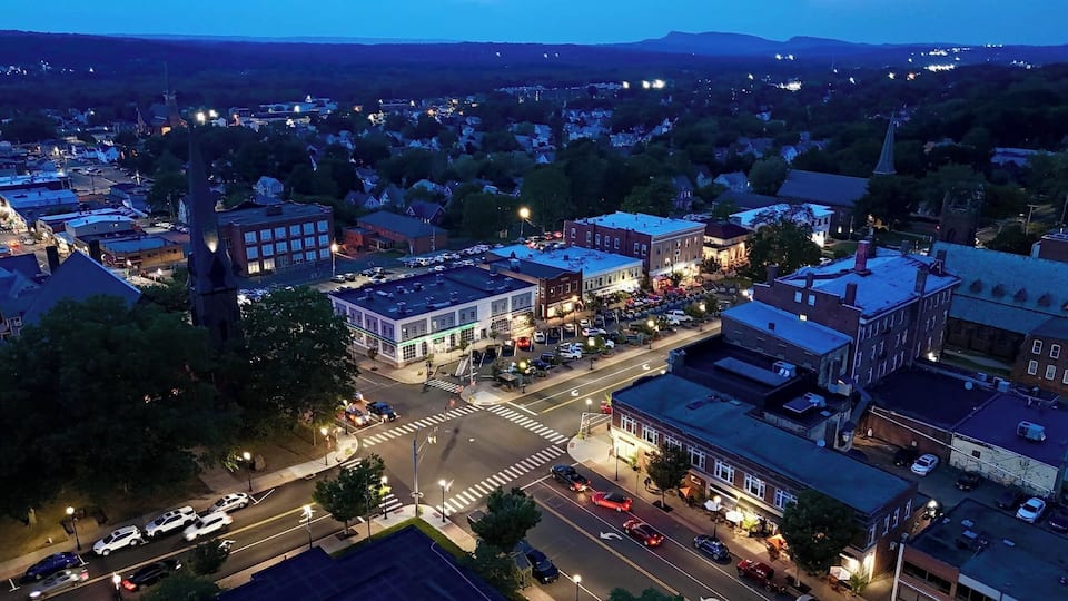 A town at dusk with illuminated streets and buildings, a prominent church steeple, and a crosswalk intersection.