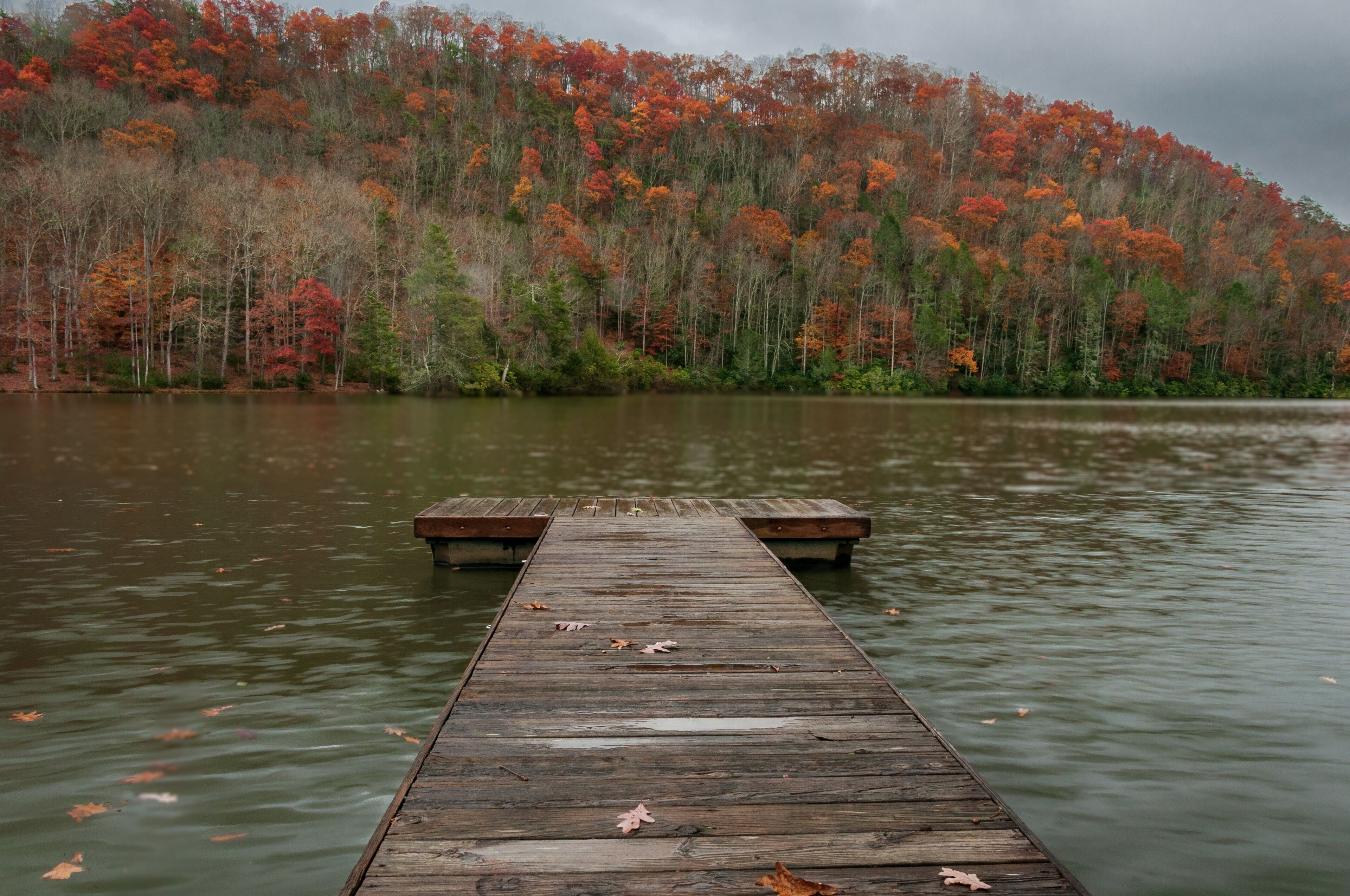 A wooden pier extending into a calm lake, surrounded by a forest with autumn-colored trees.