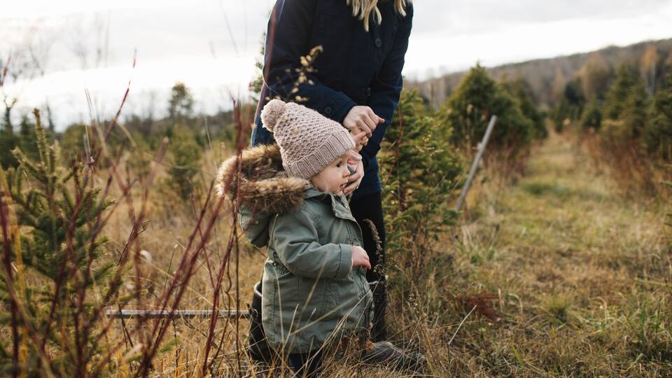 A person in a green jacket and a child in a hat, standing in a field with sparse vegetation and a forest in the background.